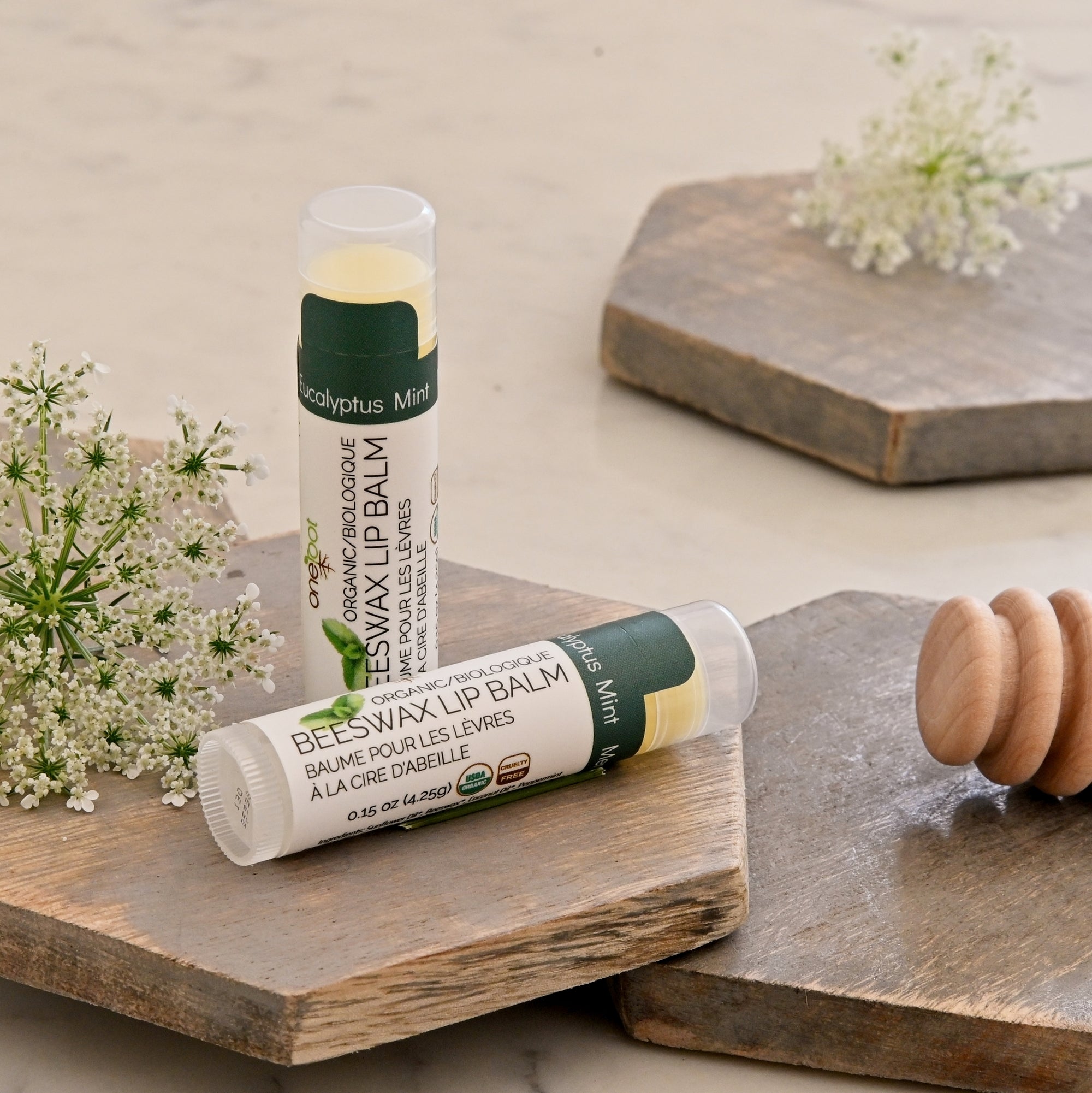 Two tubes of mint beeswax lip balm styled on rustic wooden boards with small white flowers and a wooden honey dipper on a light marble surface.