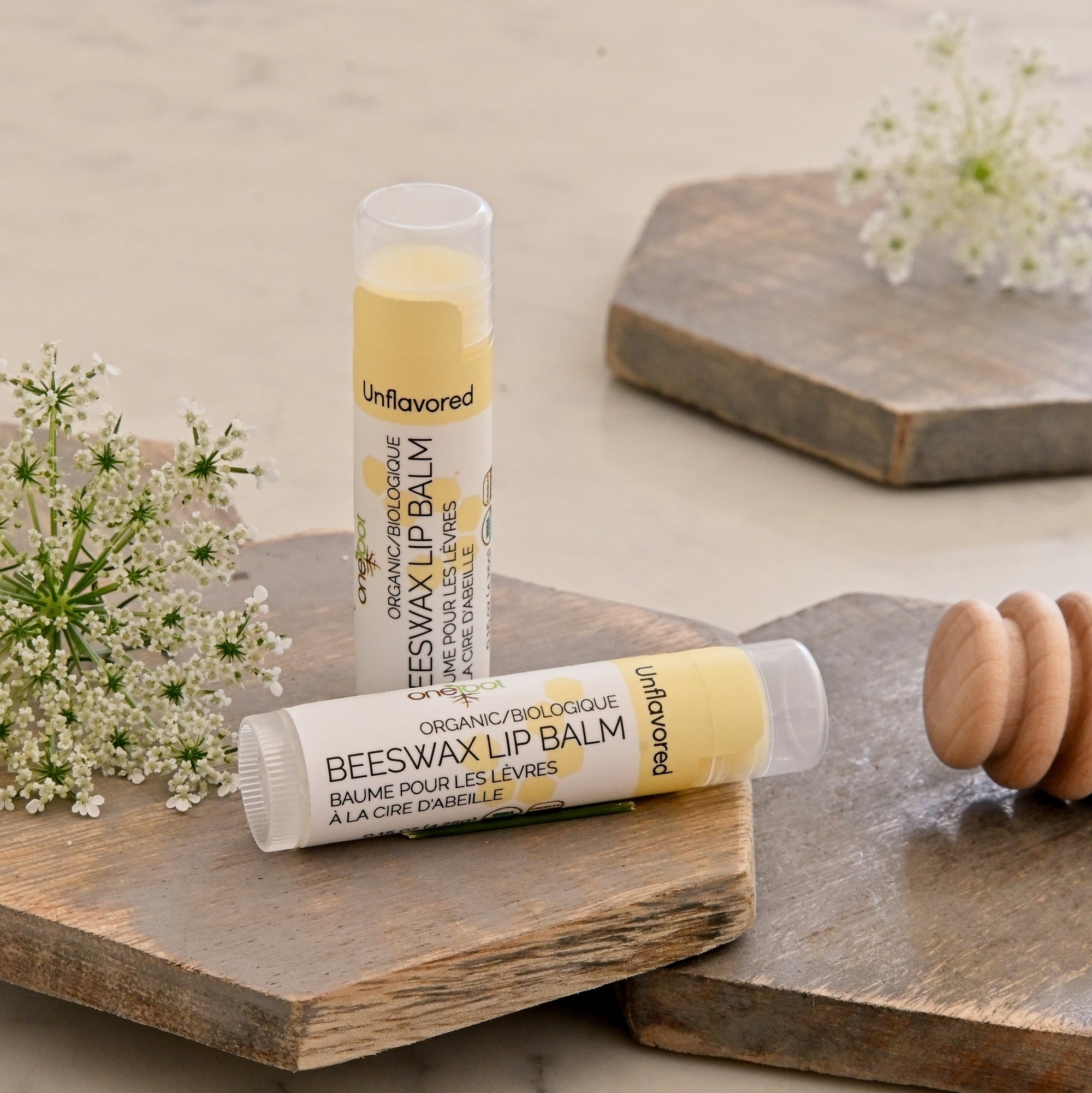 Two tubes of Unflavored Organic/Biological Beeswax Lip Balm are styled on rustic wooden boards with white flowers on a light beige surface.